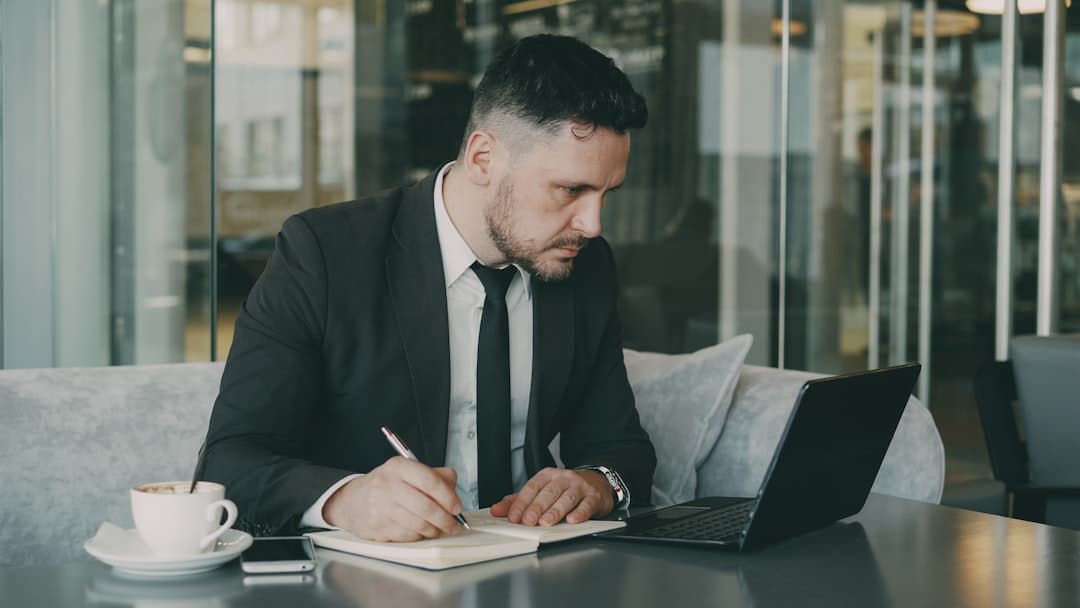 Man in suit writing in notebook at cafe table.