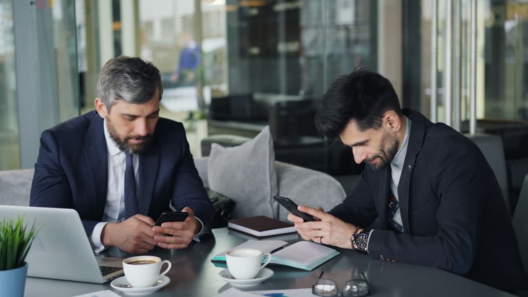 two men sitting at a table looking at their phones